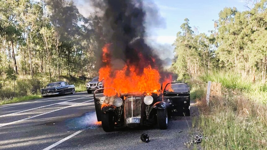 A black hot rod car on fire on the side of a road surrounded by green trees.