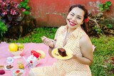 A woman eating chocolate cake and smiling, sitting on a picnic blanket with fruit and tea.