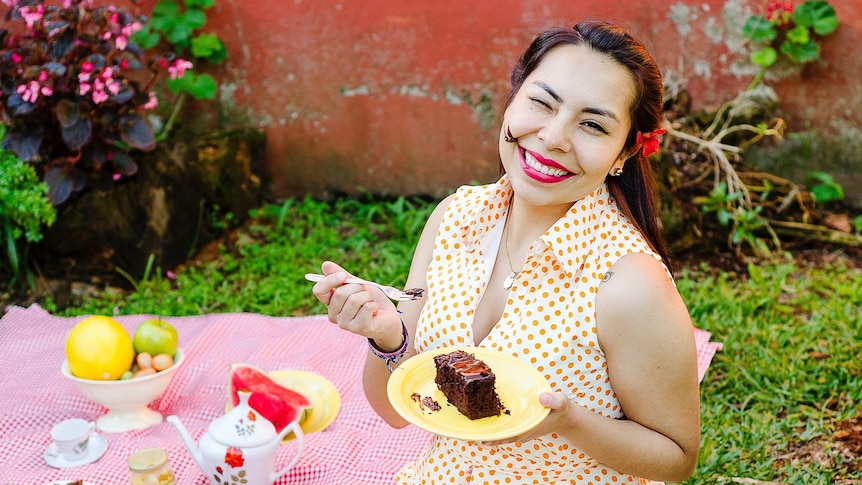 A woman eating chocolate cake and smiling, sitting on a picnic blanket with fruit and tea.