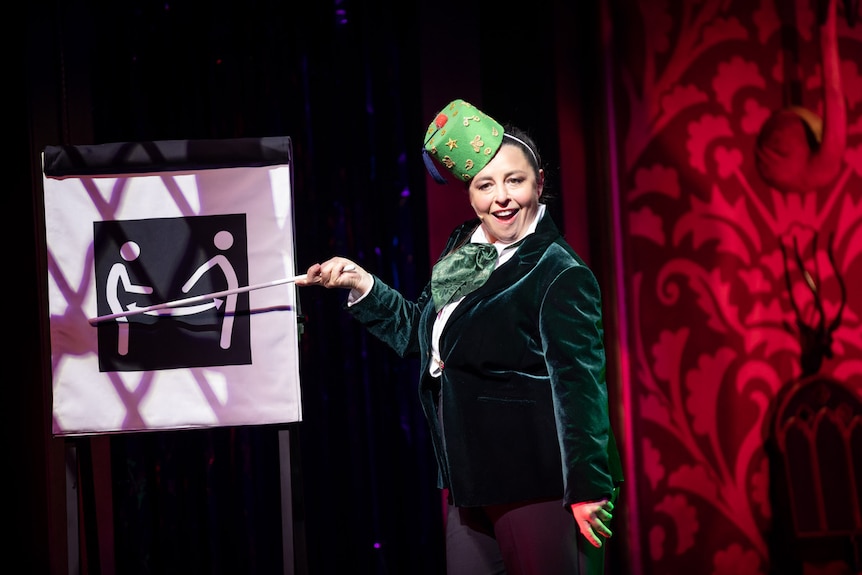 Myf Warhurst in a green velvet blazer wearing a circular hat with a tassel pointing at a diagram