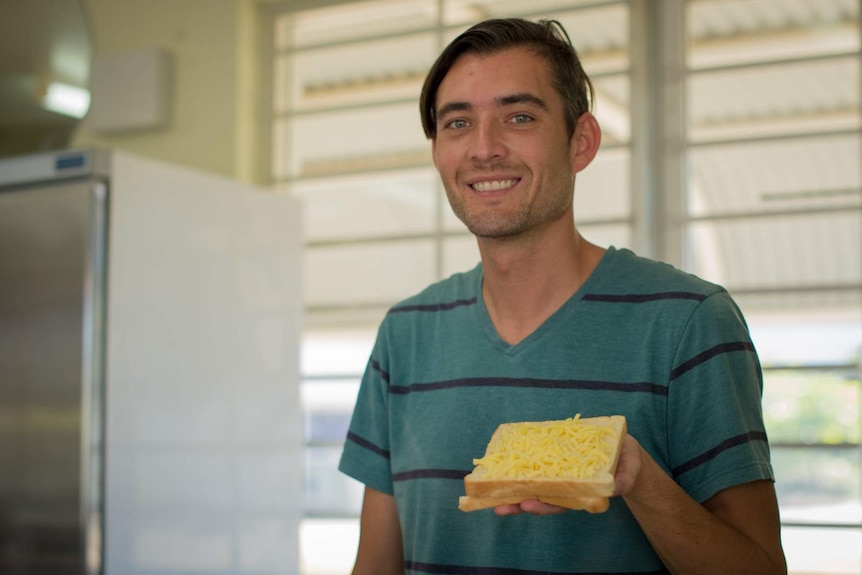 A man in a green shirt stands holding a cheese sandwich in his left hand.