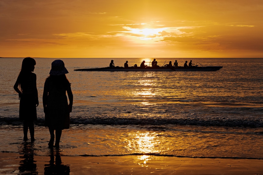 Two children stand on the beach watching a rowing boat at sunset.