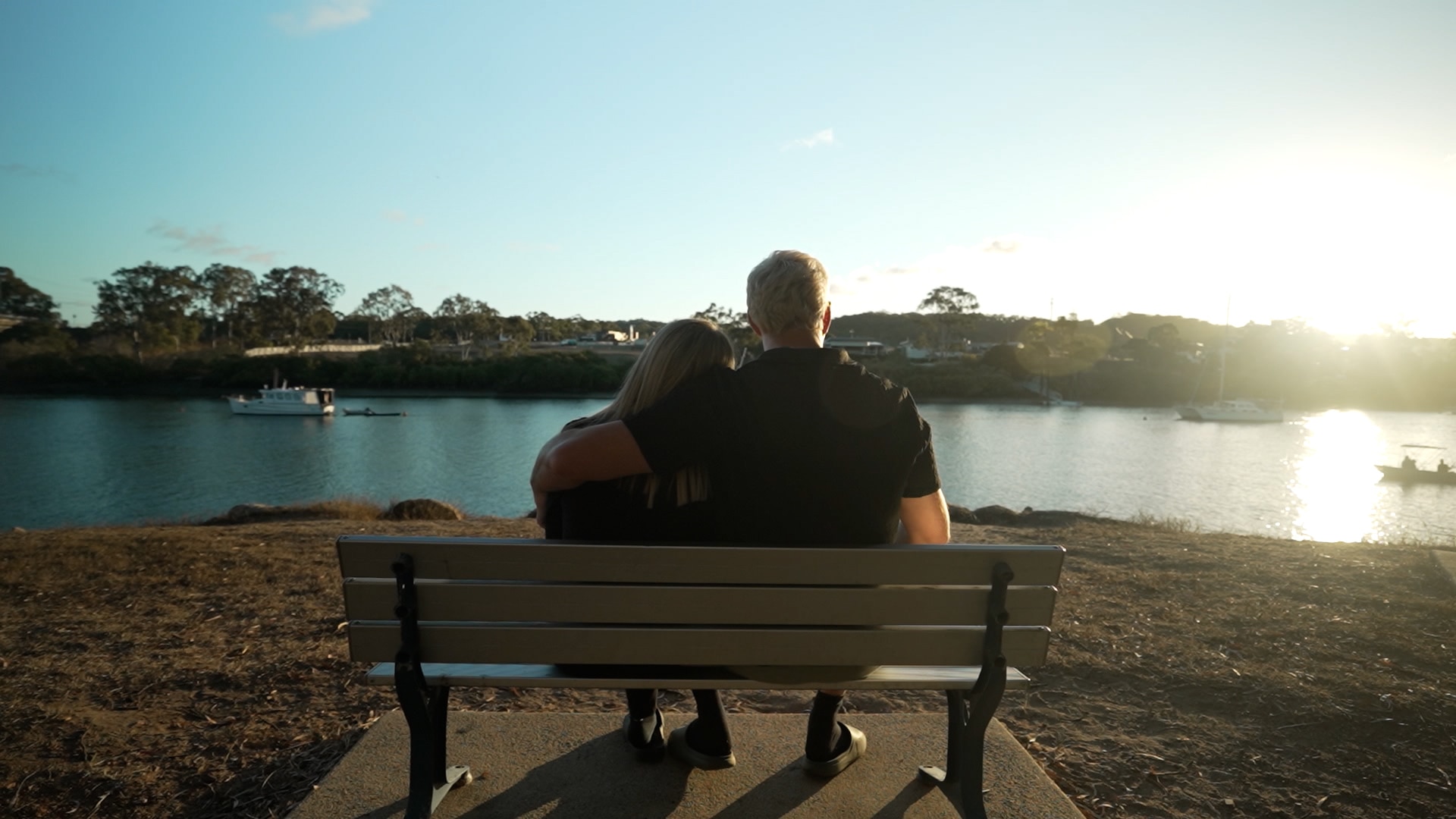 A shot from behind a bench in front of a river as two people sit shortly before sunset. The man has his arm around the woman.