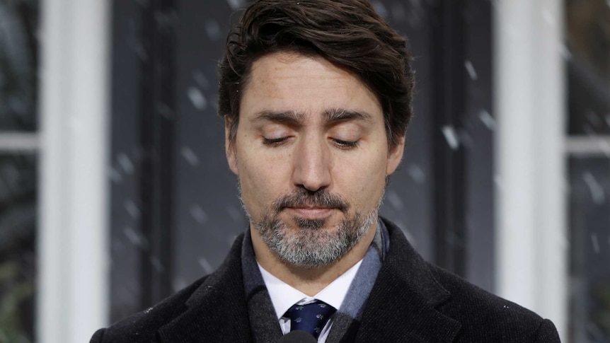 Justin Trudeau looks down with a sombre expression at a microphone with snow falling in the background