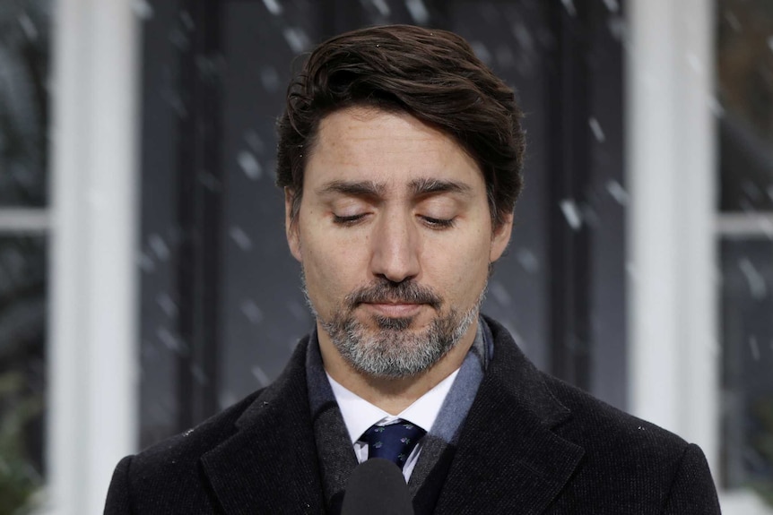 Justin Trudeau looks down with a sombre expression at a microphone with snow falling in the background