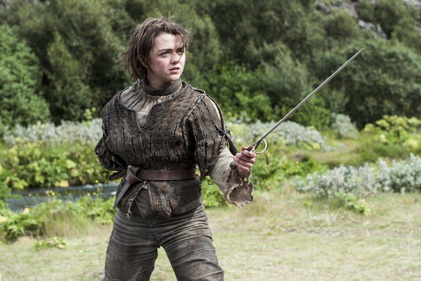 Maisie Williams brandishes a sword in front of a natural background of shrubbery.