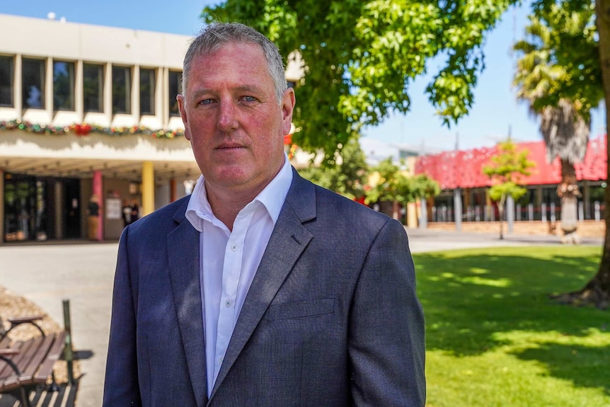 A man in a white shirt and blazer standing under a leafy tree in a park stands looking at the camera.