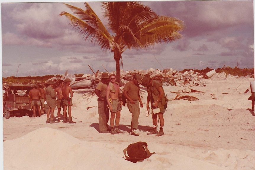 A group of soldiers line up to be checked with a Geiger counter with rubble in the background.