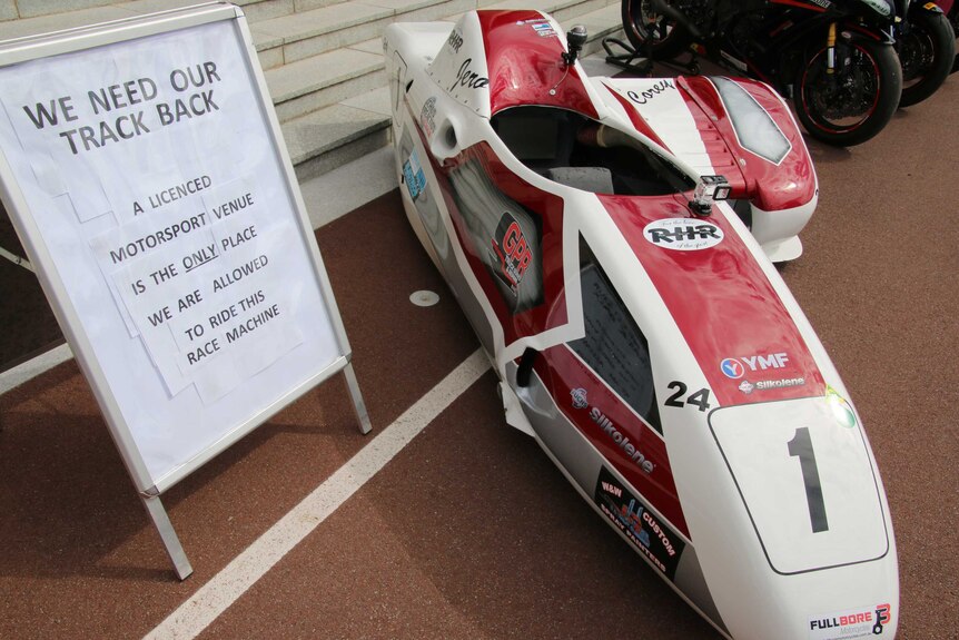 A sidecar racing bike alongside a sign calling for motorcyclists to have access to Barbagallo Raceway.