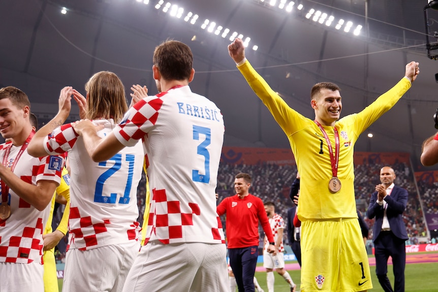 A man in a yellow goal keeper jersey with a medal with arms raised, next to men in white and red jerseys clapping