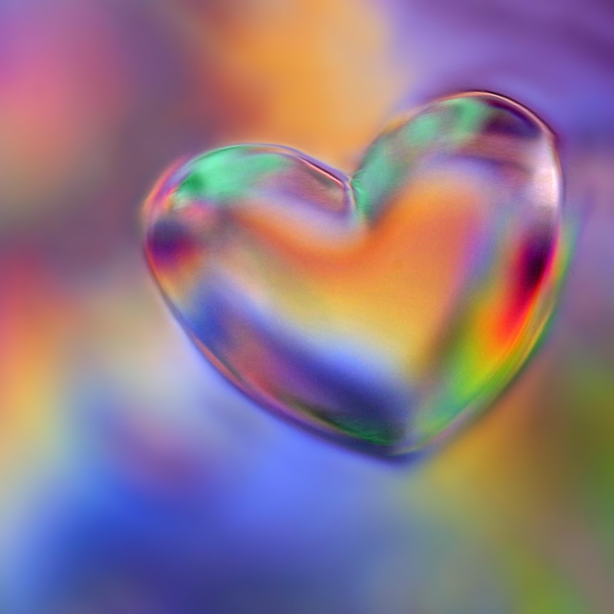 Close up of a water drop in the shape of a heart with a holographic rainbow inside and behind it. 