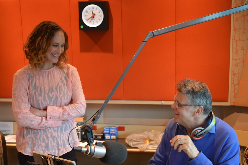 Dina standing talking to audio engineer sitting at radio studio desk with microphone in foreground.
