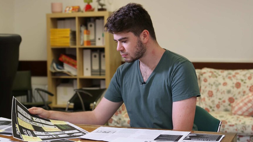 A young man sits at a desk wearing a dark green shirt looking at pages.
