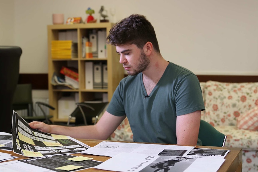 A young man sits at a desk wearing a dark green shirt looking at pages.