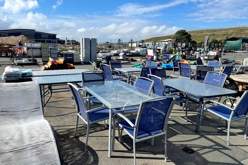 Heaps of tables, chairs and other outdoor furniture, blue sky and green grass hill in background.