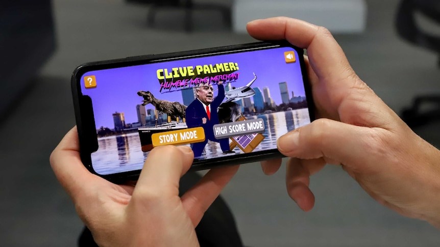 Hands hold an iphone, with a Clive Palmer app on screen, showing a cartoon Palmer wearing a suit next to a dinosaur and plane.
