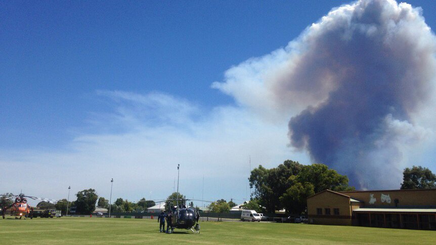 Two helicopters sit on a sporting field in Harvey as a big cloud of smoke rises into the sky from a nearby fire.