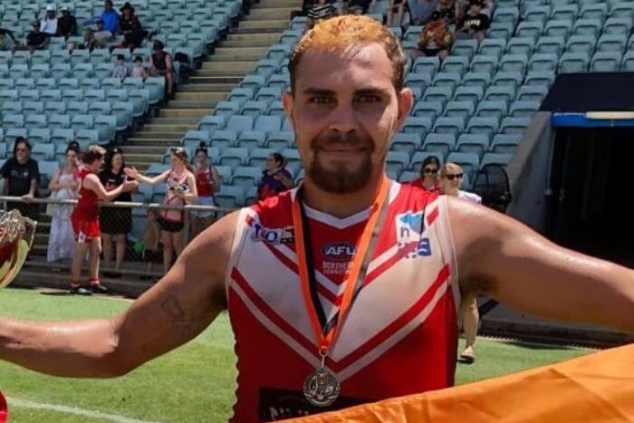 a happy footballer after a game holding a premiership flag and wearing a medal