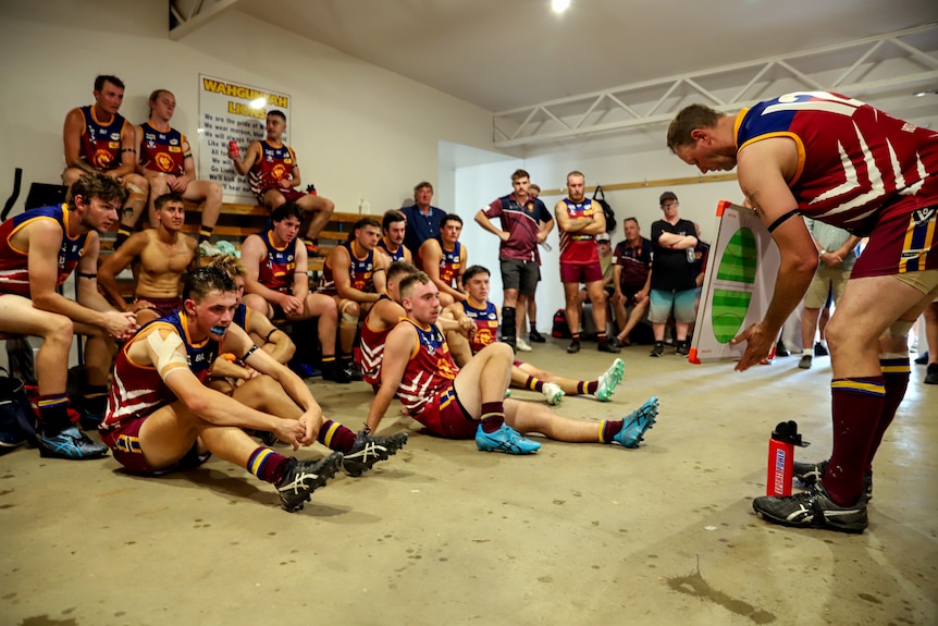 Man wearing football uniform holds magnet board in front of two dozen other footballers inside concreted change rooms