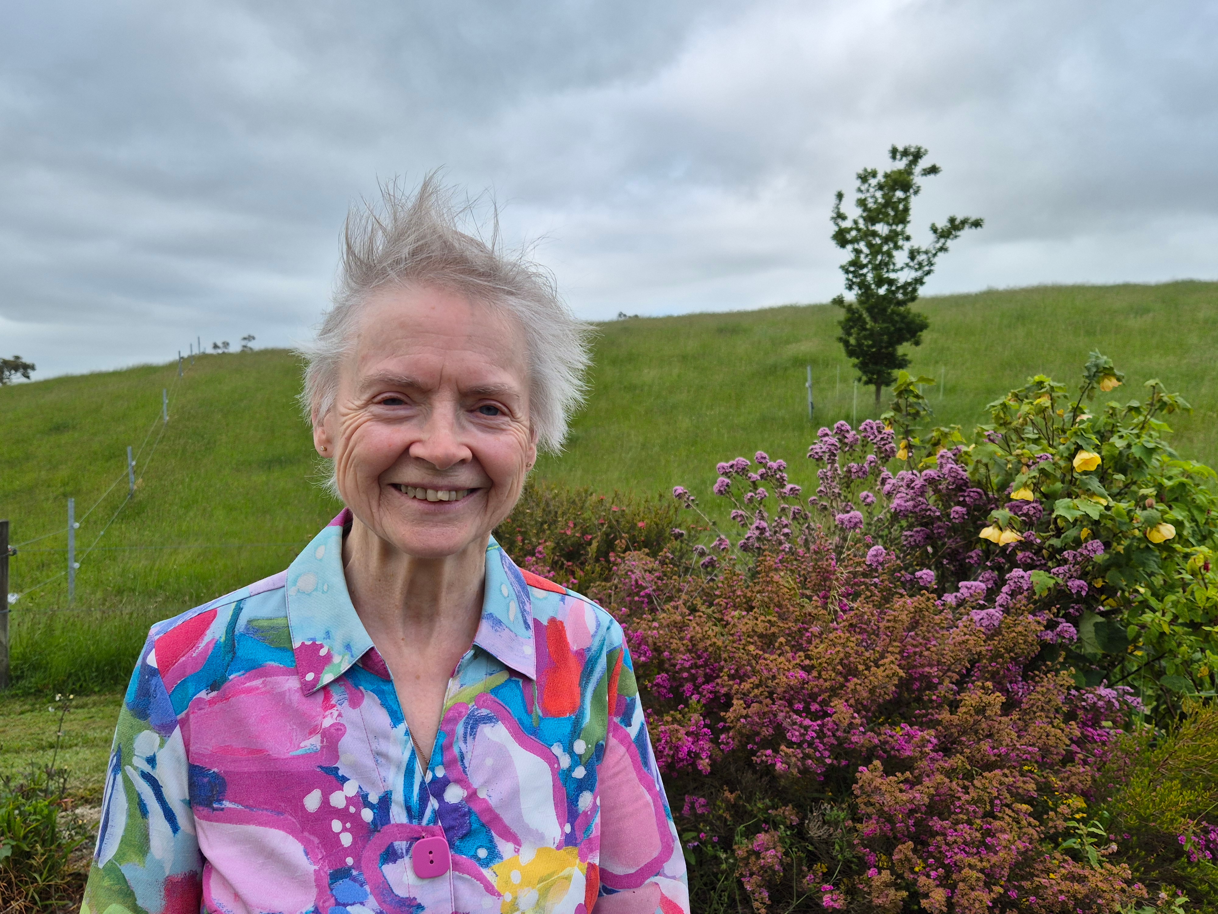 Uma senhora idosa com uma camisa colorida sorri para a câmera, em frente a uma colina verde e um jardim iluminado.