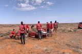 People in red shirts around a small solar car and desert landscape