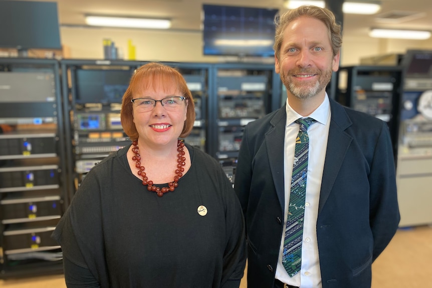 A man with a beard in a suit smiles at the camera and a woman with red hair and a red necklace smiles