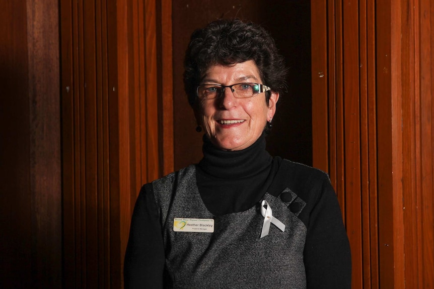 A woman standing against a wooden wall wearing a white ribbon and with a name tag saying Heather Blackley