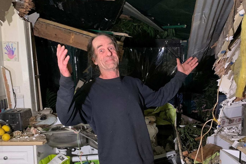 A man stands in his destroyed kitchen with his hands up.