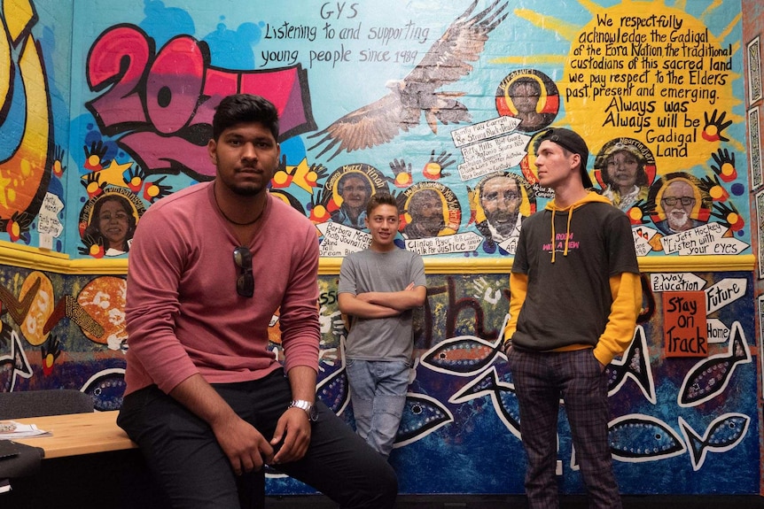 Three students stand in front of a mural at the Glebe Youth Service