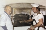 Two men smile in front of an old oven while cooking in it. 