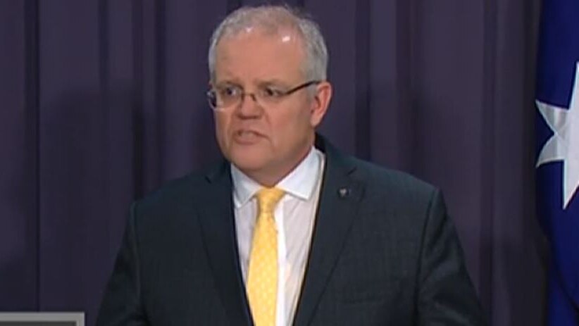 The Prime Minister of Australia in a dark suit and yellow tie, next to the national flag.