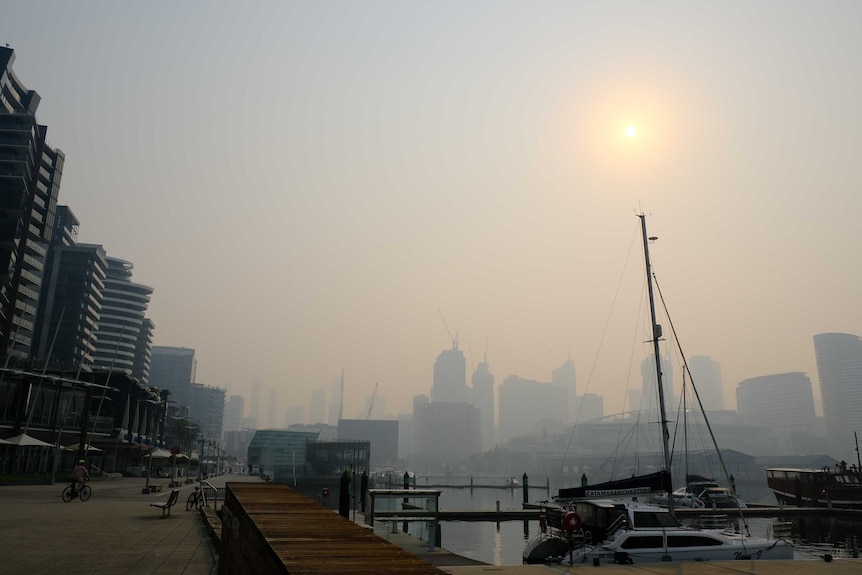 A boat sits on the water in front of a smoky Melbourne skyline, where the zone is a pale yellow behind the smoke.