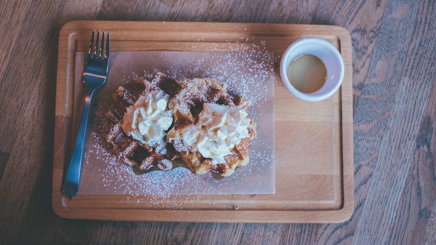 Wooden serving board with a plate of waffles covered in cream and coffee in a white mug.