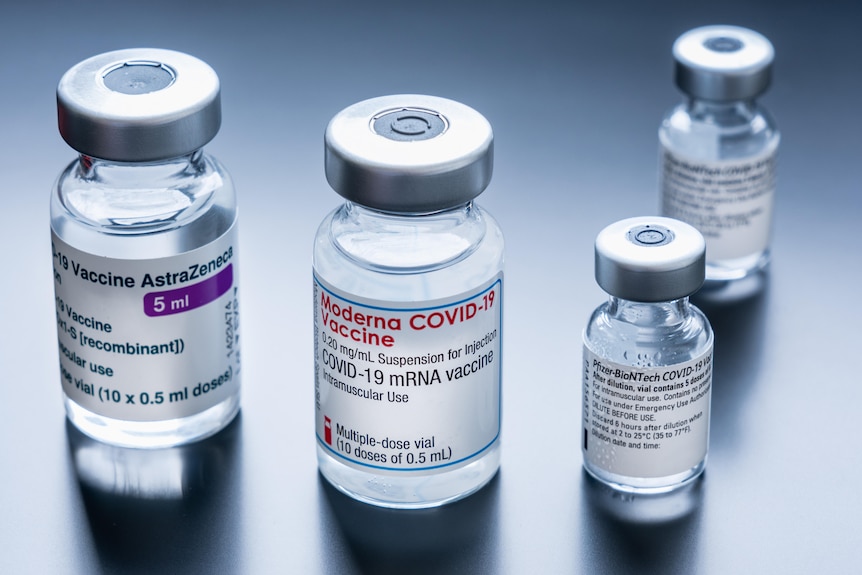 One vial with Moderna, one with AstraZeneca and two with Pfizer are arranged on a plain gray background.