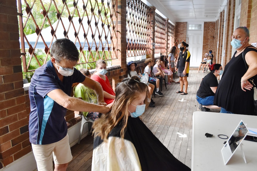 A hairdresser cuts a girl's long hair.  There's a line of people waiting behind them