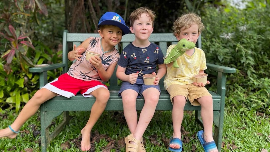 Three little boys sit on a bench outdoors.