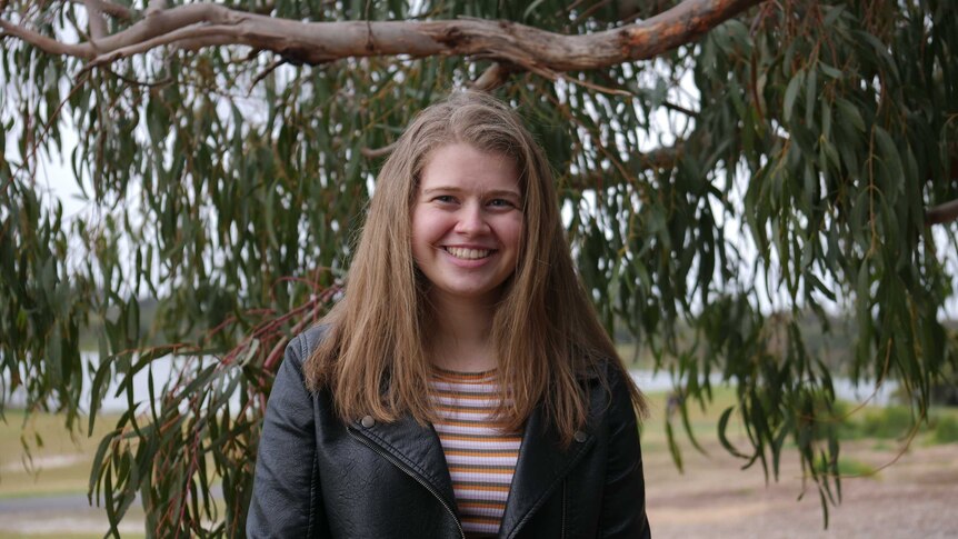 A blonde haired person with striped top and leather jacket, stands in front of gum tree, smiling
