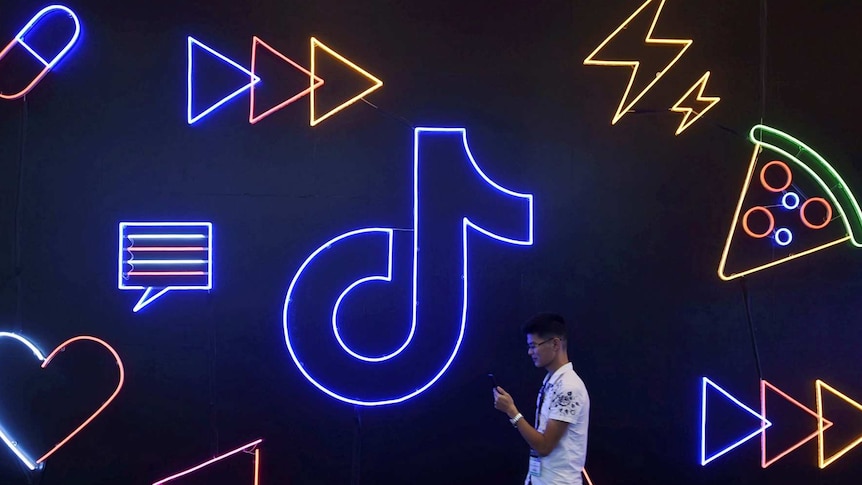A man walks past a neon sign in the shape of the TikTok logo, while looking at his phone