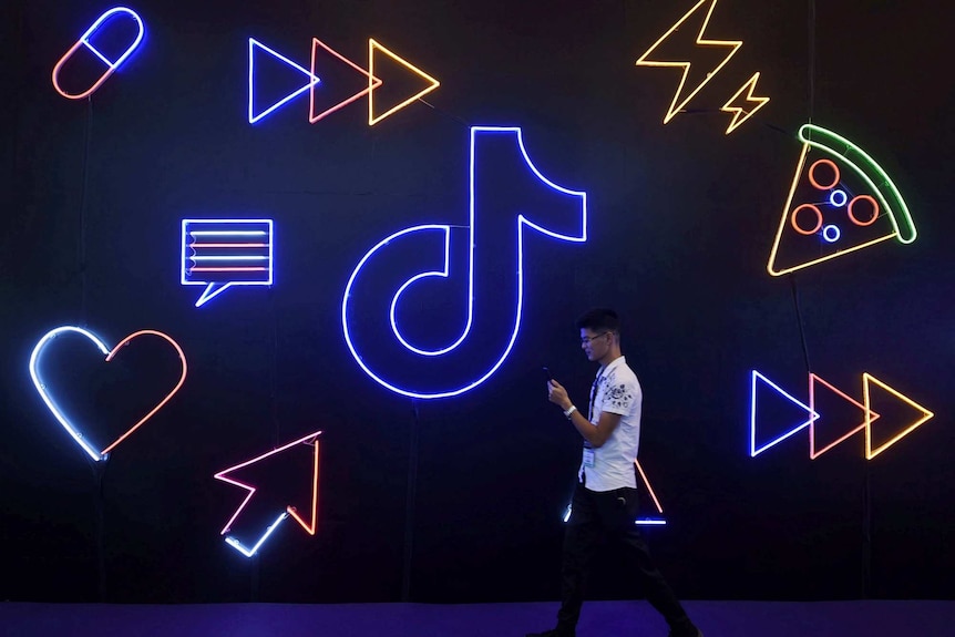 一名男子一边看手机，一边走过一个TikTok标志形状的霓虹灯招牌