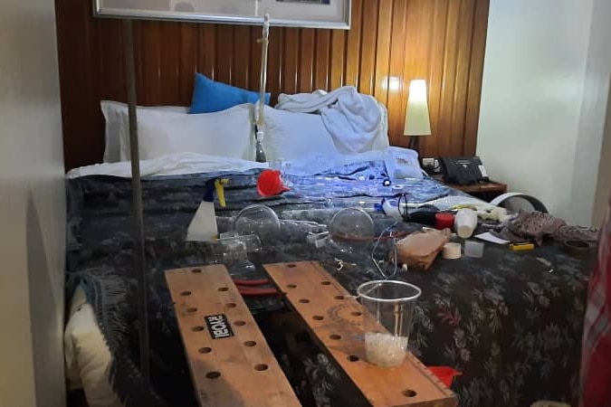 Equipment scattered across a bed in a hotel room. There is a cup with a white substance in it, in the foreground.