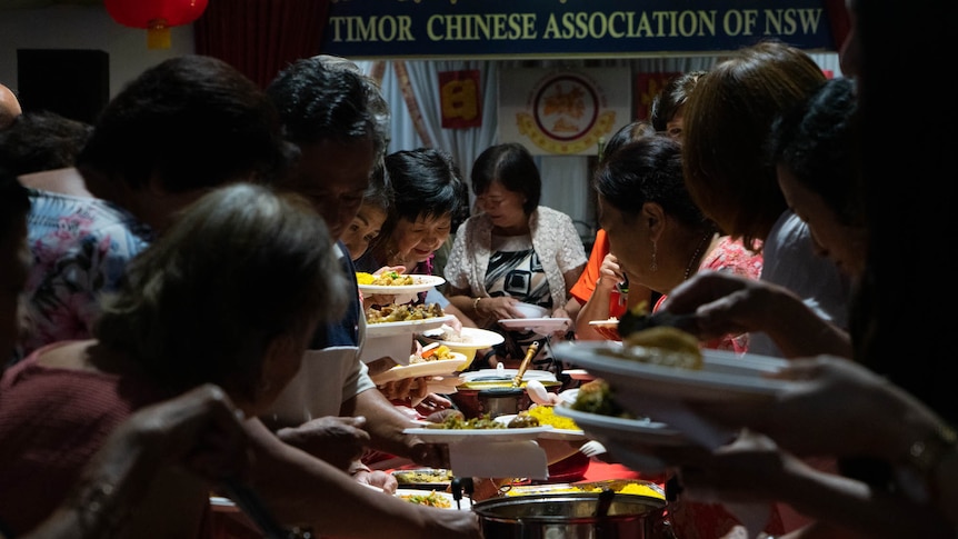A crowd of people lean in with cutlery and crockery to a table, lit up, covered in bowls and plates of food.
