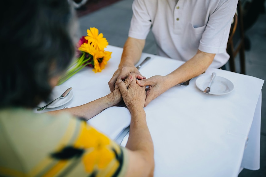 A close up of an elderly woman and man holding hands at a restauarant there is a bunch of flowers on the table next to them 