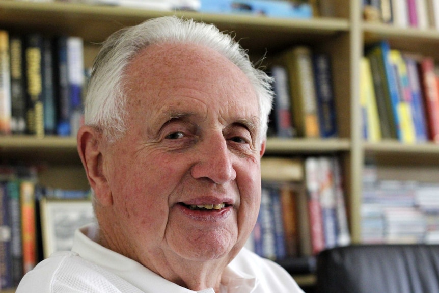 An elderly man with white/grey hair sitting in front of a bookshelf.