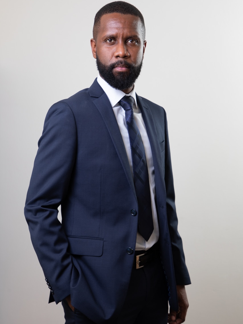 A man in a blue suit with a carefully groomed beard looks seriously at the camera. 