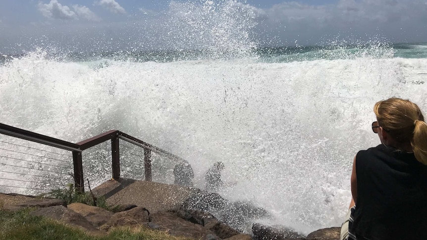 A big wave crashes onto people sitting on stairs down to the beach at Snapper Rocks.