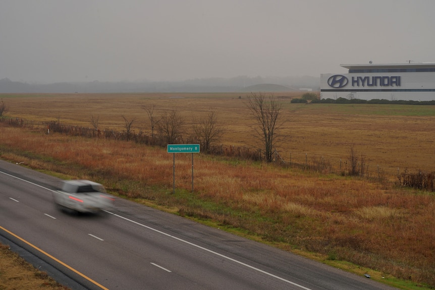 a car drives down a road next to a paddock next to a factory with a Hyundai sign on it.