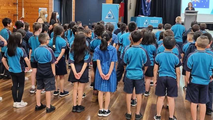 a group of primary school students standing in a school hall with their backs to the camera