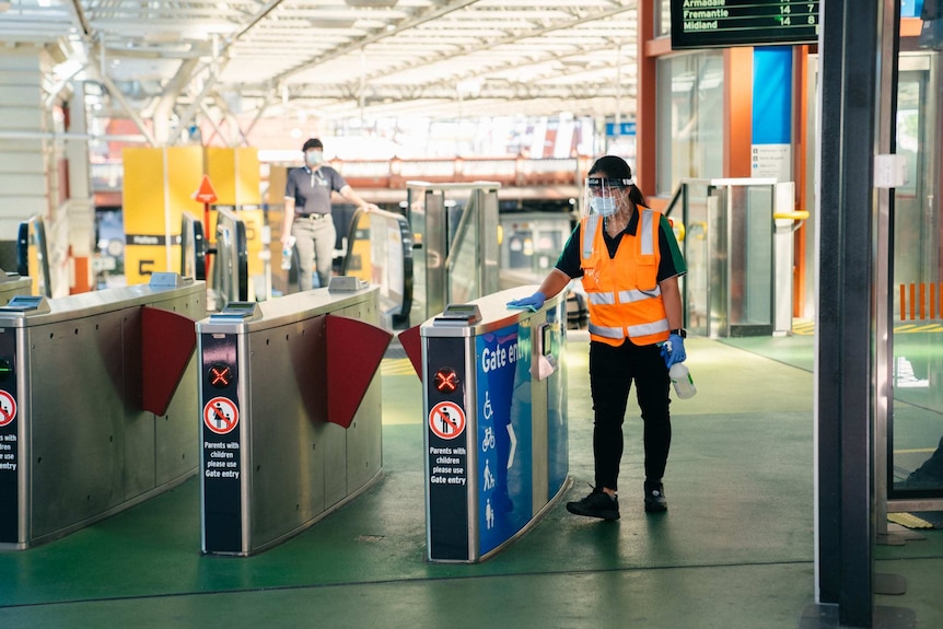 A lady in high vis and a full face mask wipes down gate entry at perth train station