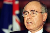 John Howard, with bushy dark eyebrows and his signature glasses, stands in front of an Australian flag.