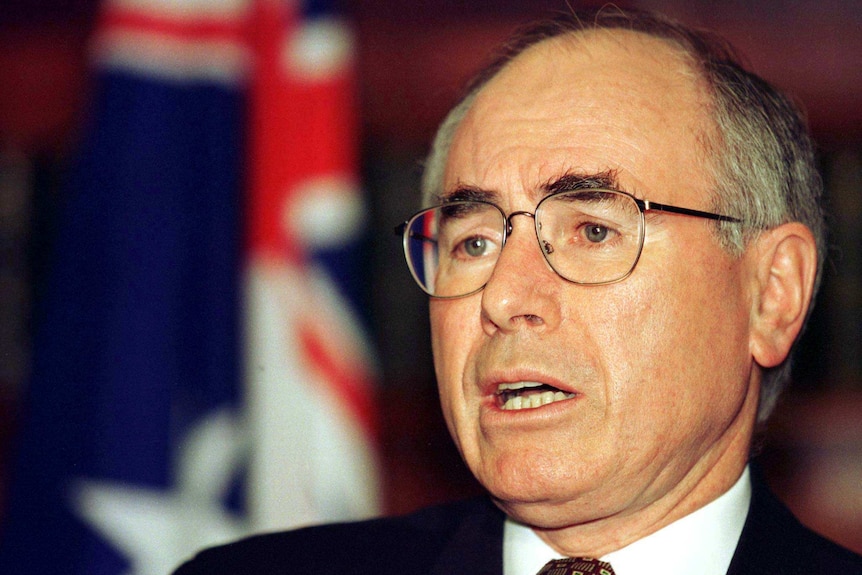 John Howard, with bushy dark eyebrows and his signature glasses, stands in front of an Australian flag.